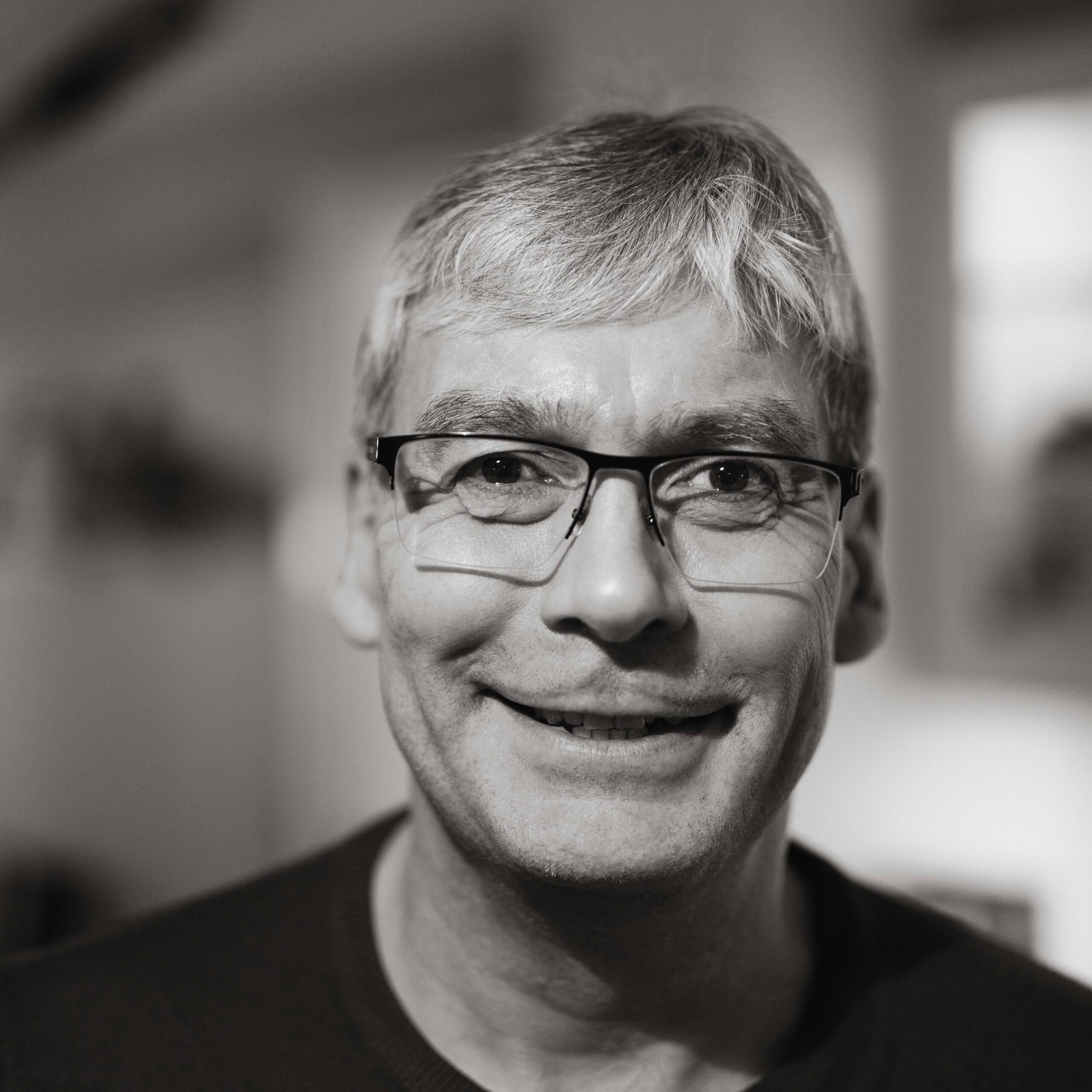 Close-up black and white portrait of a smiling man wearing glasses indoors.