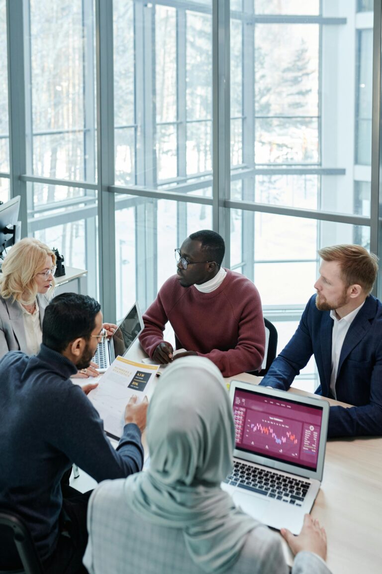A diverse business team discusses reports and statistics around a wooden table with laptops indoors.
